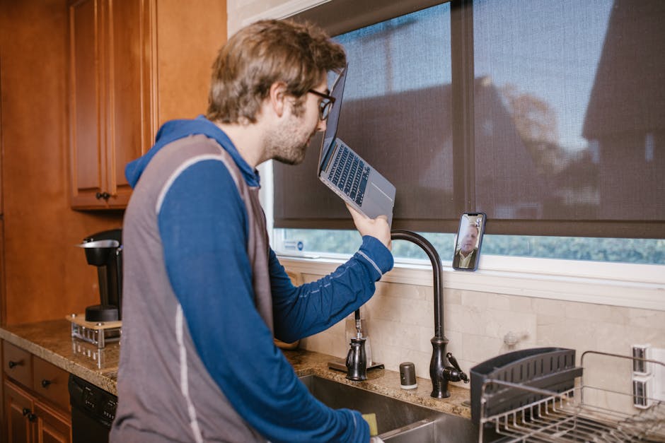 A man in a kitchen making a video call using a laptop and smartphone.